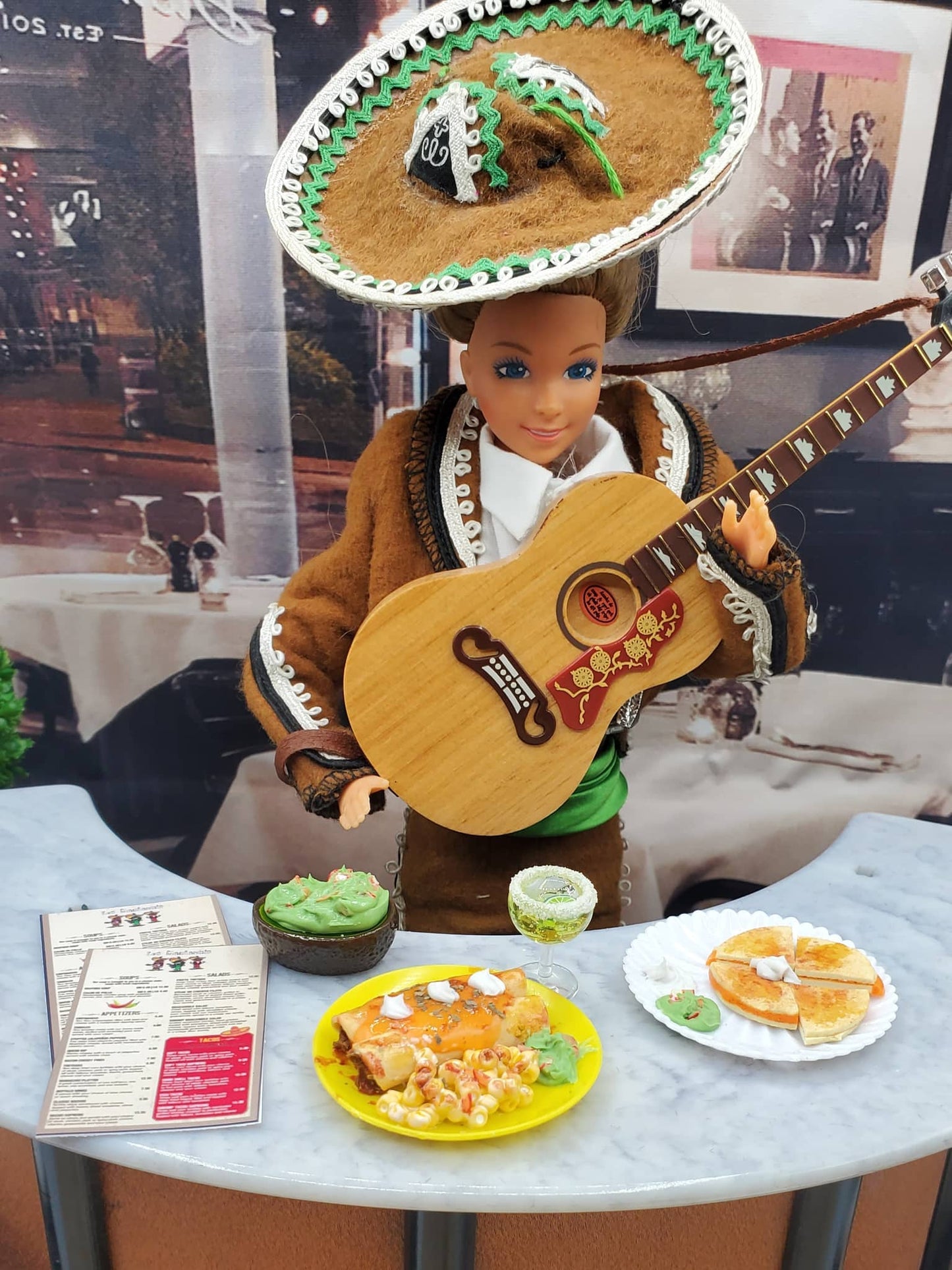 Doll in traditional Mexican attire with a guitar, sombrero, and food on a table.