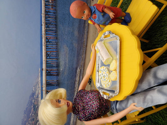 Barbie doll serving lemon sponge rolls for dolls on a yellow table with a child doll by the beach