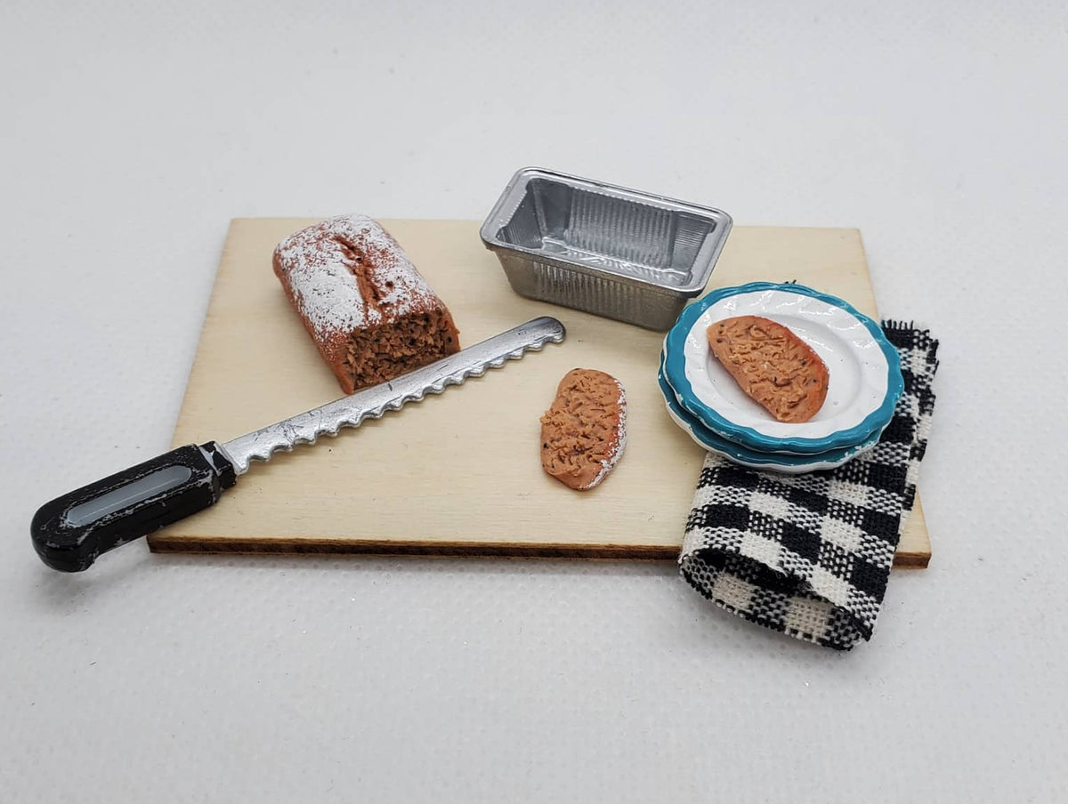 Miniature banana bread for dolls with a serving plate, knife, and baking pan on a wooden board
