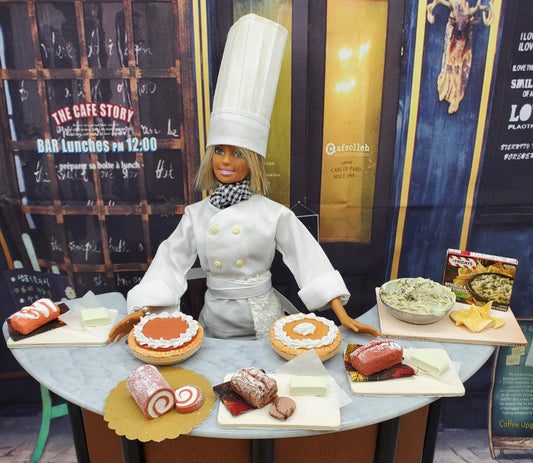 Miniature banana loaf for dolls displayed with other doll-sized desserts on a table with a doll chef in white uniform