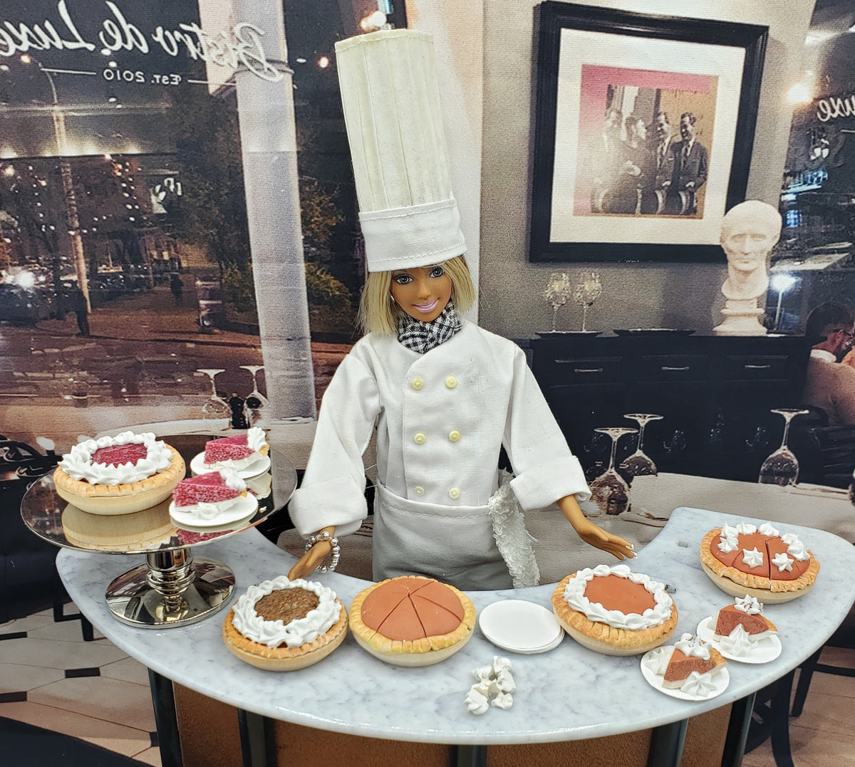 Miniature cherry pie for dolls displayed with other miniature desserts by Chef Gina's Mini Food behind a chef doll in white uniform