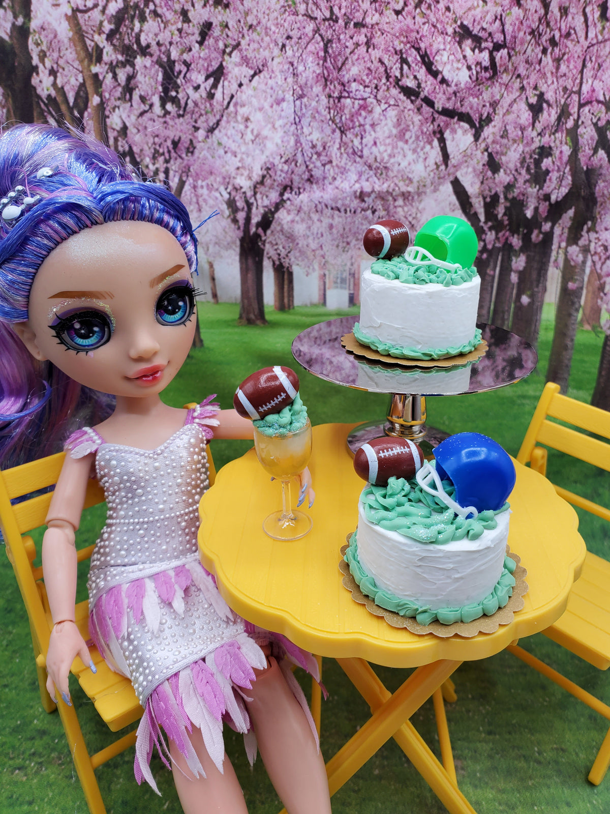 Chef Gina's miniature football cake decorated with football and helmet toppers on a yellow table with a doll in a garden setting