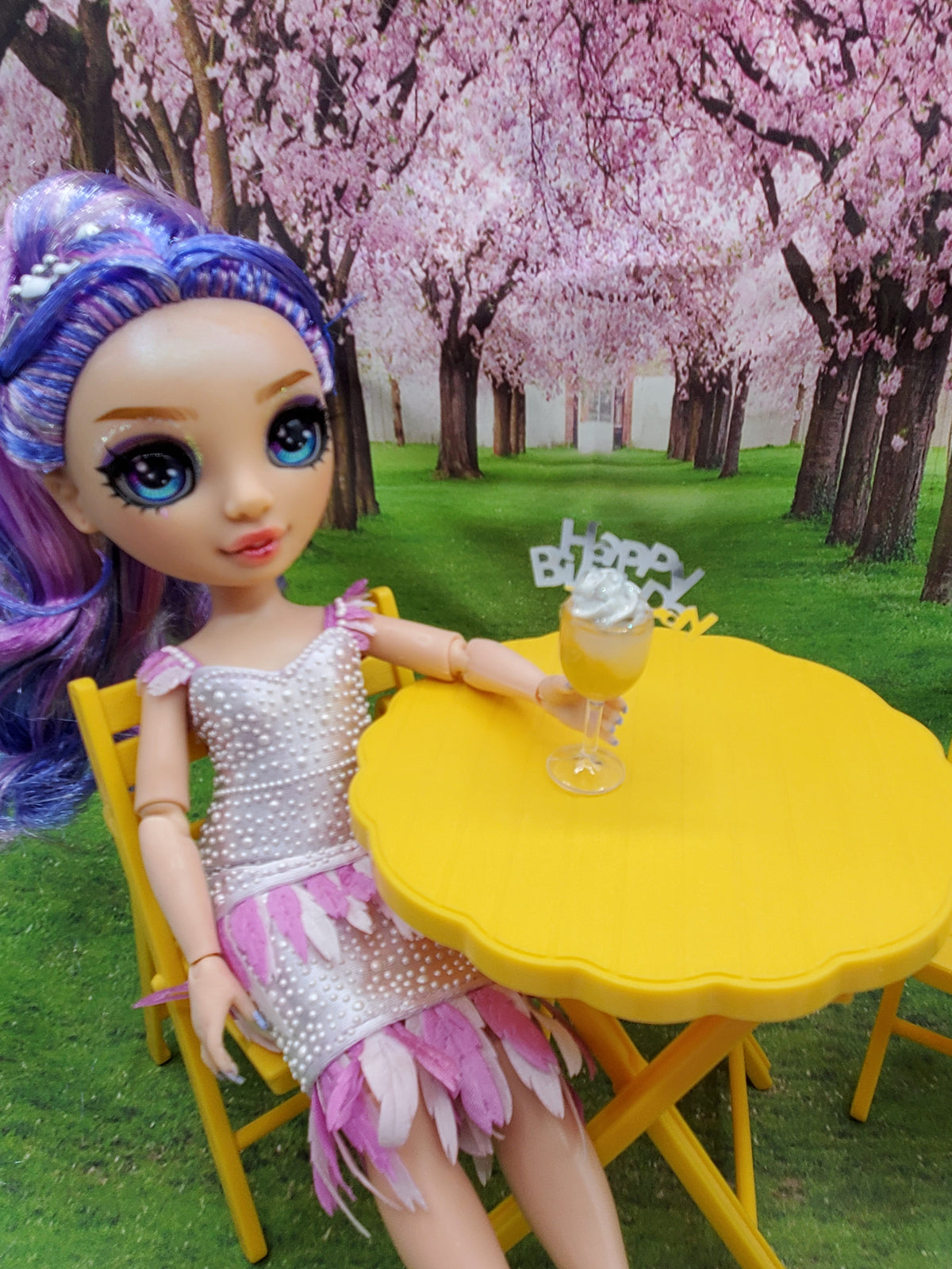 Miniature happy birthday cocktail on yellow table next to doll with purple hair in outdoor cherry blossom setting