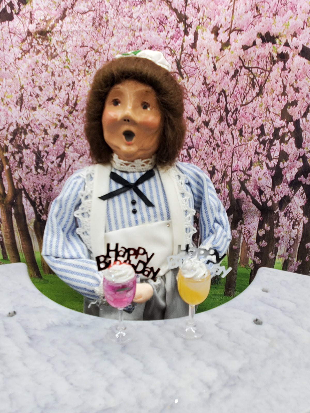 Miniature happy birthday cocktail drinks in pink and yellow held by a doll wearing a blue striped shirt and apron