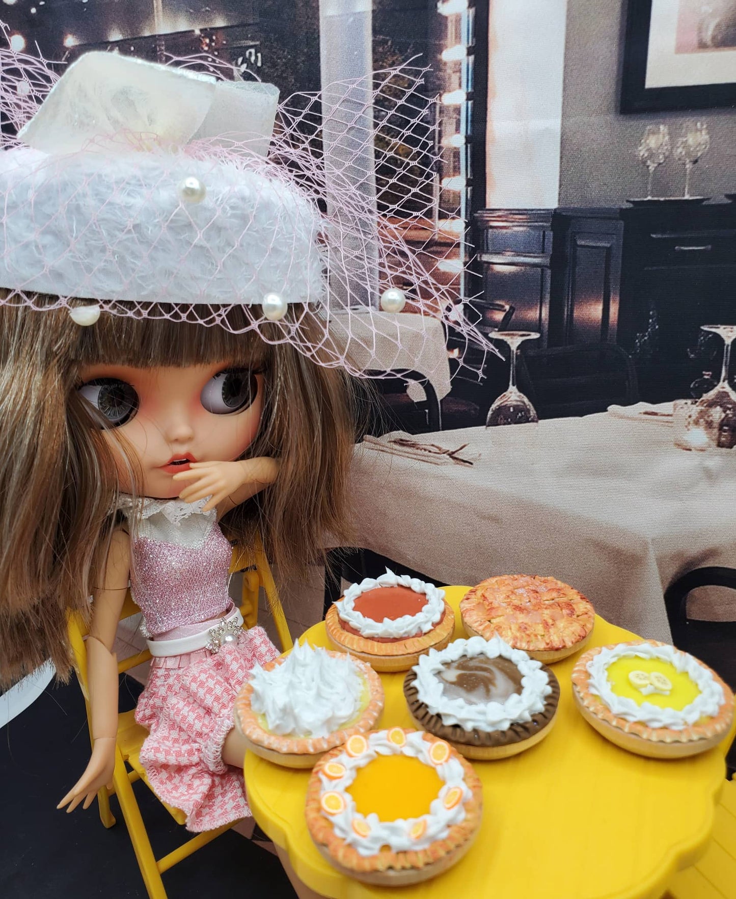 Miniature lemon meringue pie and assorted pies displayed on a yellow table with a doll in a pink dress and white hat