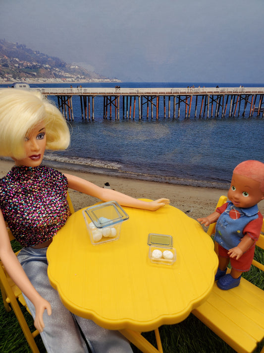 Barbie doll and child doll at a beach table displaying miniature rum balls for dolls