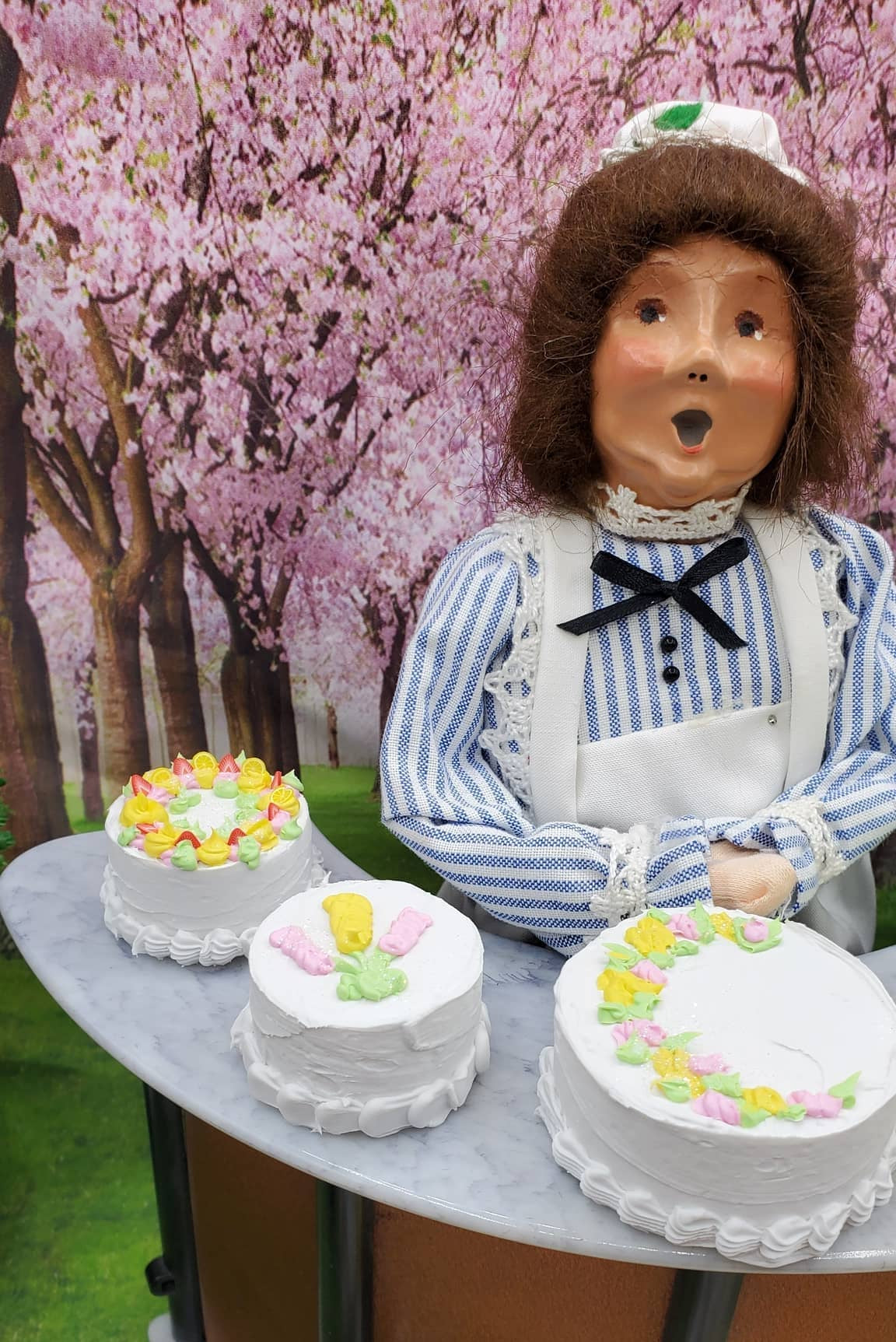 A vintage doll or figurine dressed in a blue-and-white striped nurse outfit with a white apron and black bow, posed behind a table with three decorated cakes against a pink cherry blossom backdrop.