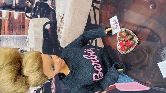 Barbie doll wearing a black outfit with 'Barbie' written on it, sitting on a chair.