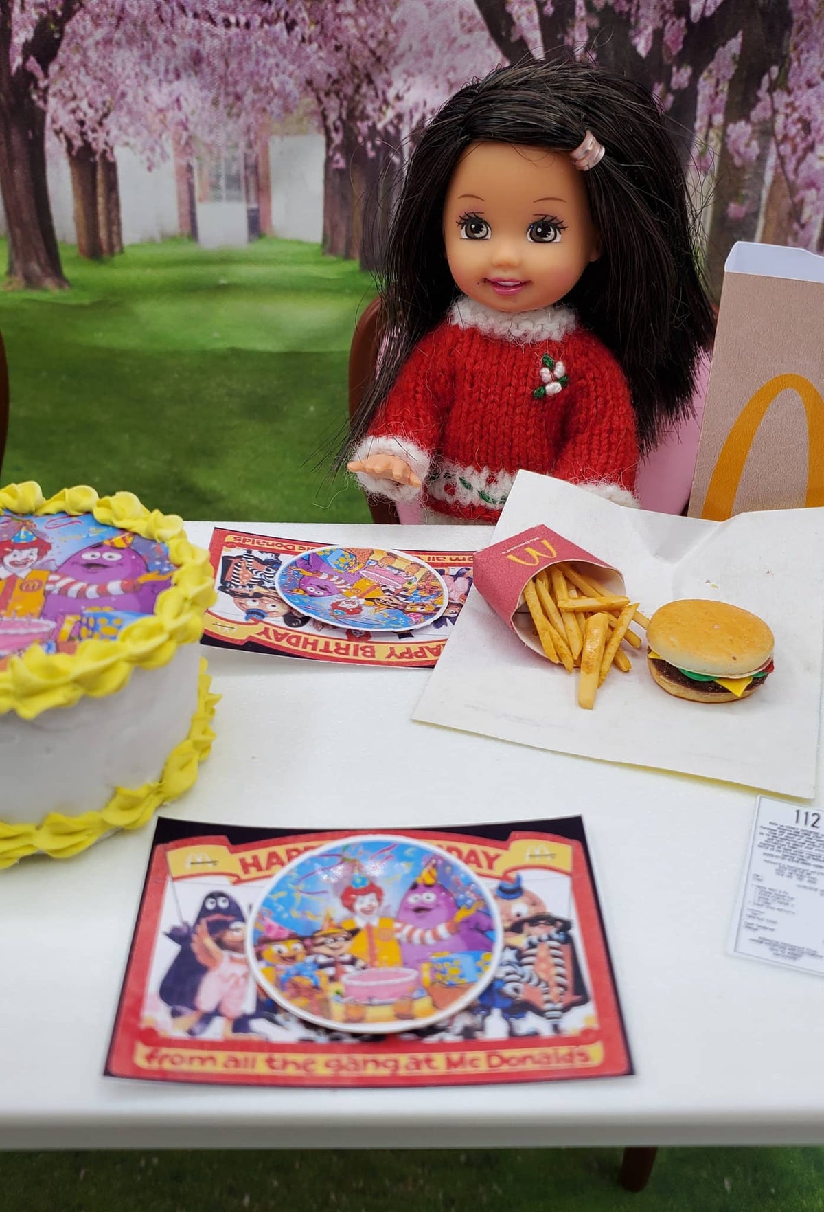 Toy doll with McDonald's Happy Meal items and a birthday card on a table outdoors.
