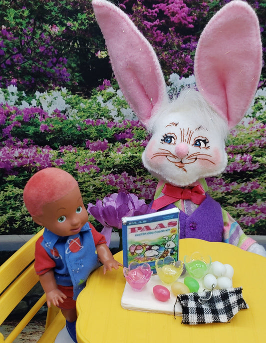 Fashion doll egg decorating kit featuring a small doll and a bunny with decorating supplies on a yellow table outdoors