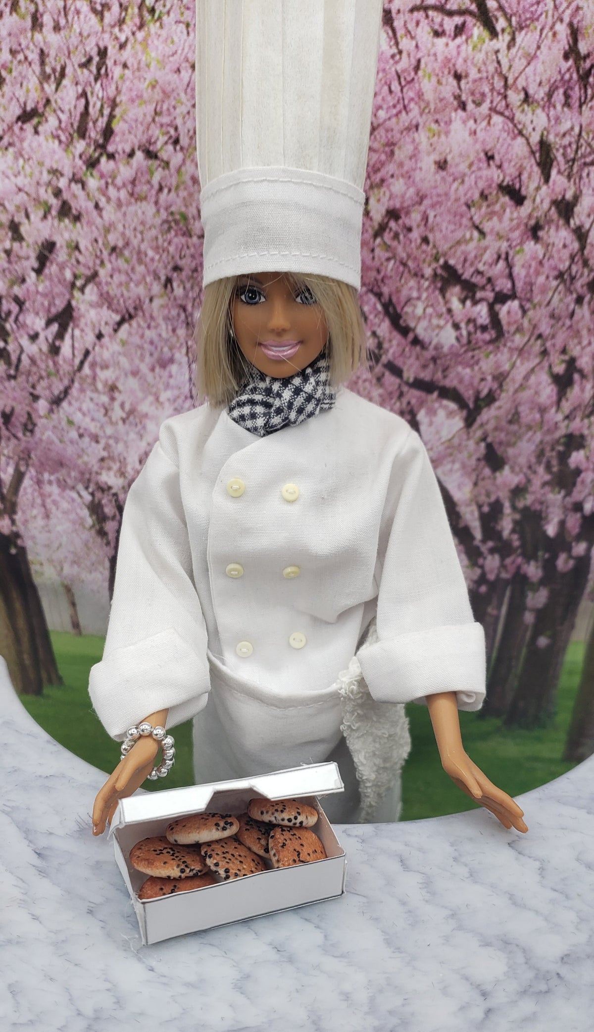 Chef Gina's doll dressed as a chef holding a box of miniature chocolate chip cookies on a marble surface