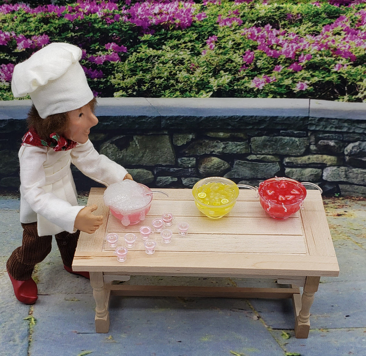 Miniature dollhouse party punch set with three colorful bowls and tiny glasses beside chef doll figure