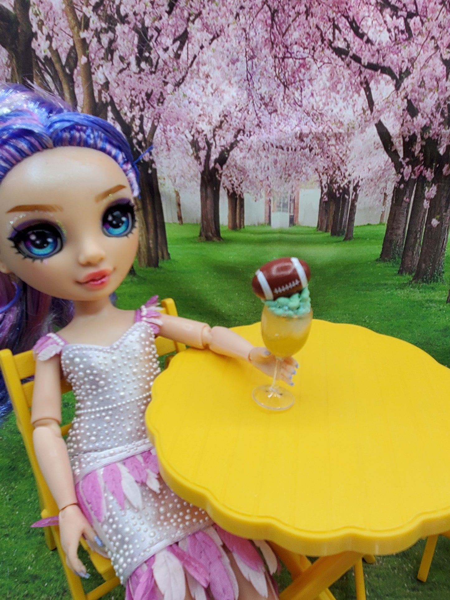 1:6 scale doll football drinks displayed on a yellow table with a doll seated in a garden setting