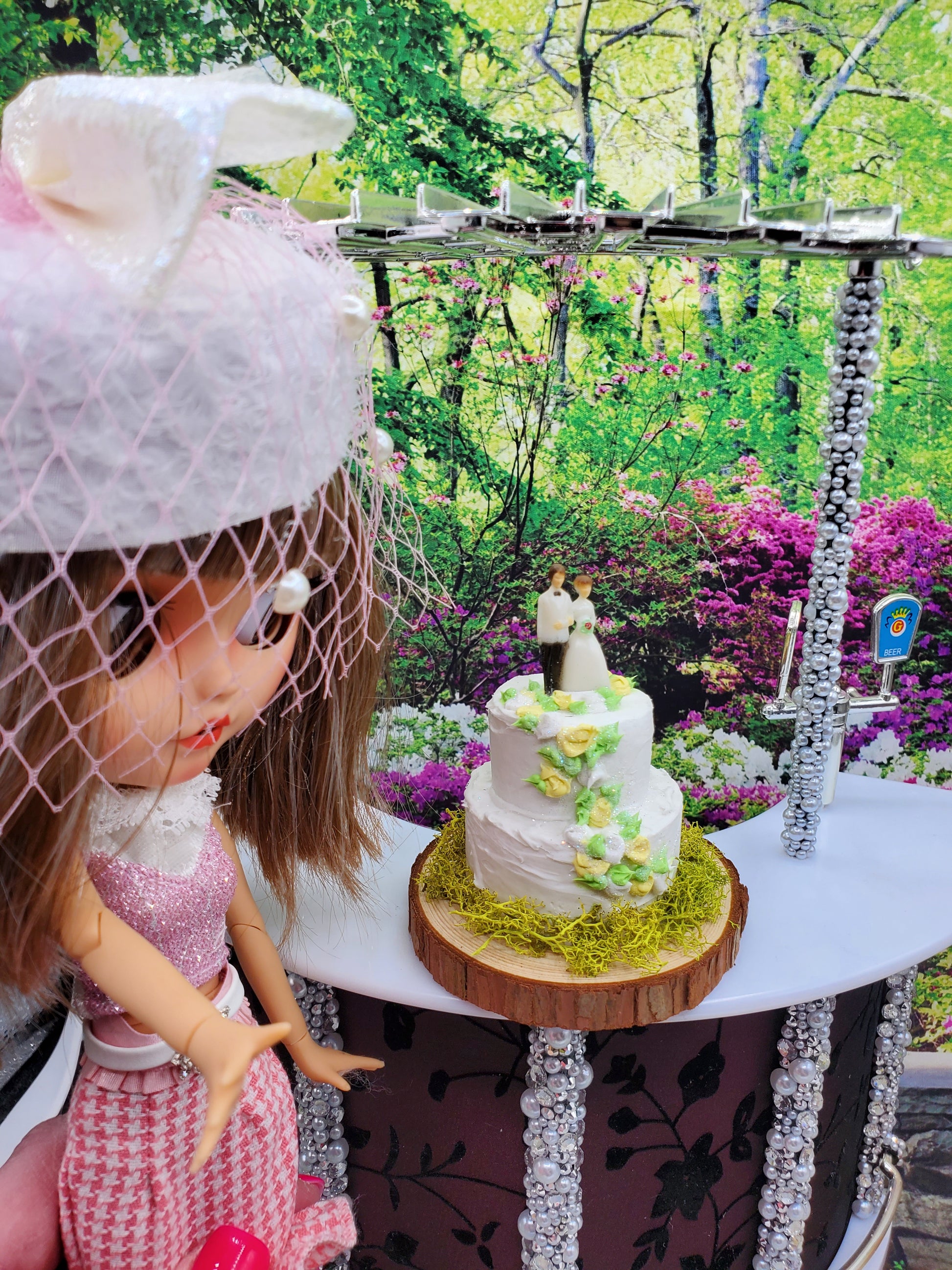 Doll next to a 1:6 scale wedding cake with white frosting and floral decorations on a wooden stand