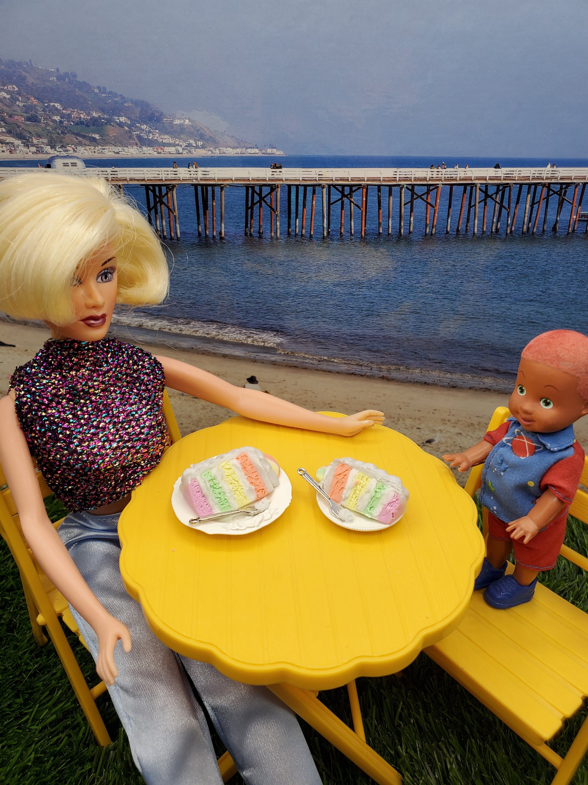 Fashion doll enjoying a sherbet cake with a child at a yellow table by the beach