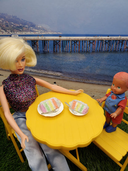 Fashion doll enjoying a sherbet cake with a child at a yellow table by the beach