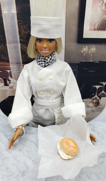 Chef Gina's miniature bagel with cream cheese held by a doll dressed as a chef on marble surface