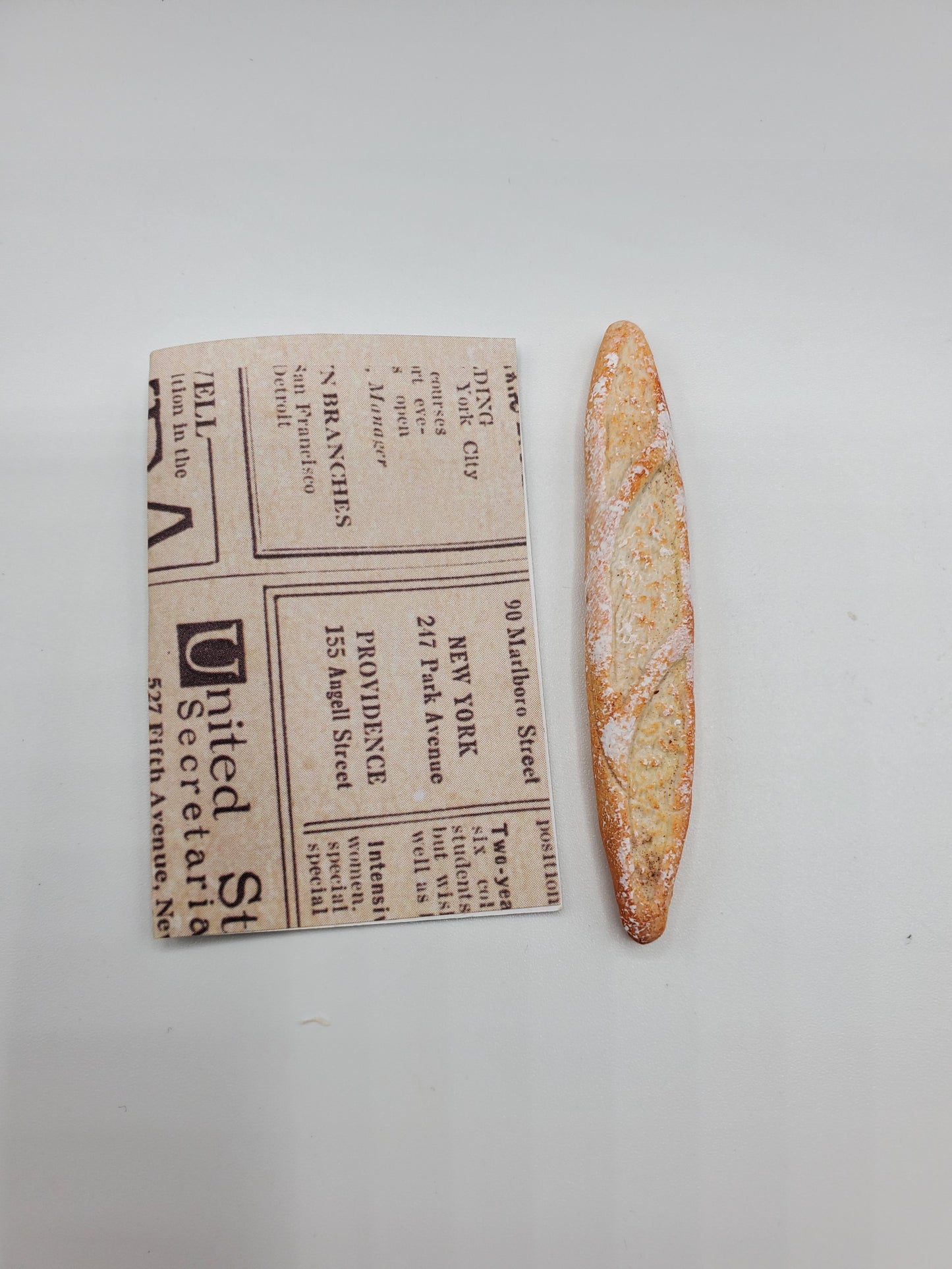 Chef Gina's miniature baguette bread next to a small folded newspaper for size comparison on a white background
