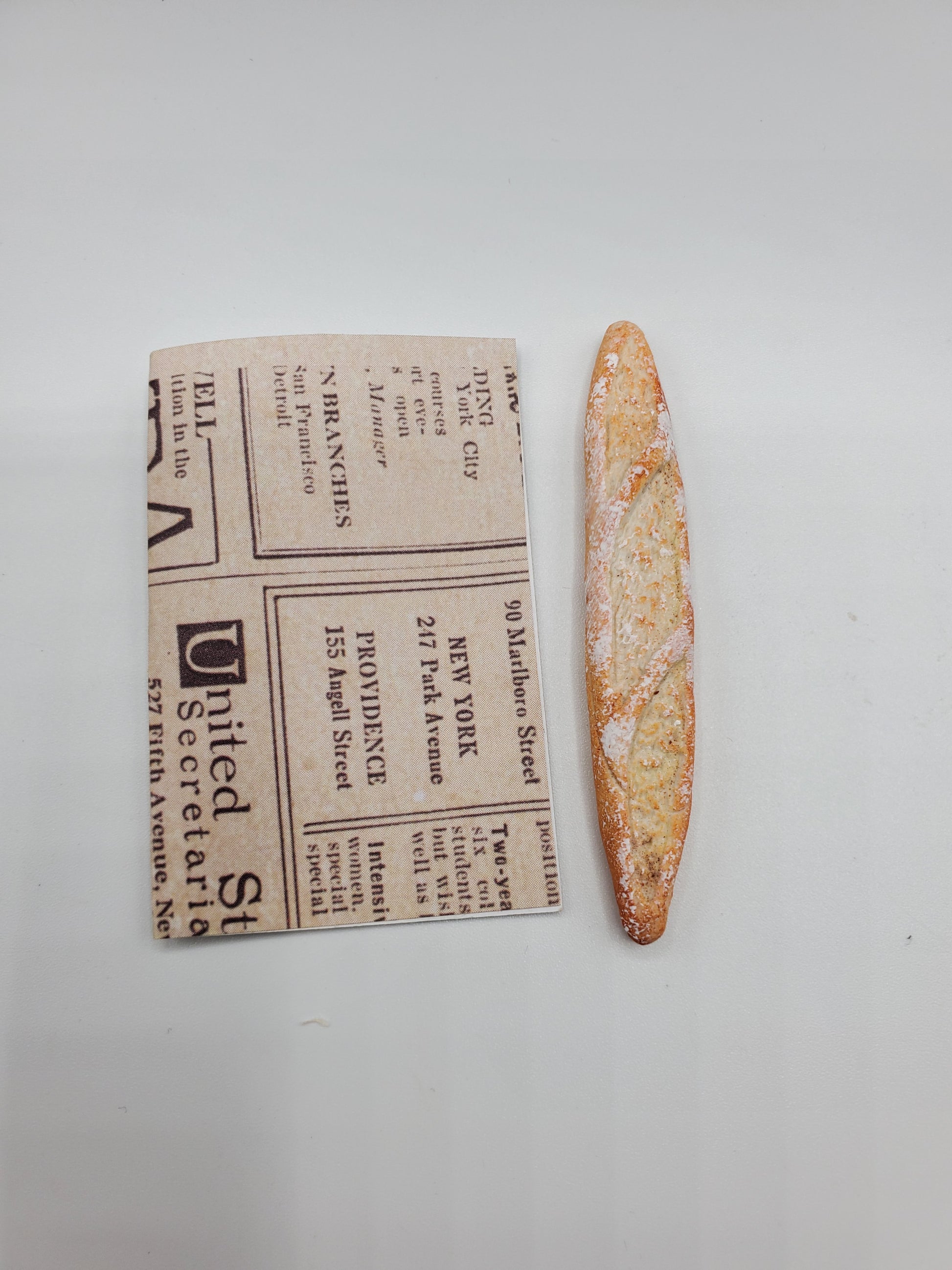 Chef Gina's miniature baguette bread next to a small folded newspaper for size comparison on a white background