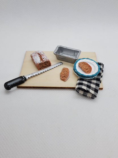Miniature banana bread for dolls with a serving plate, knife, and baking pan on a wooden board