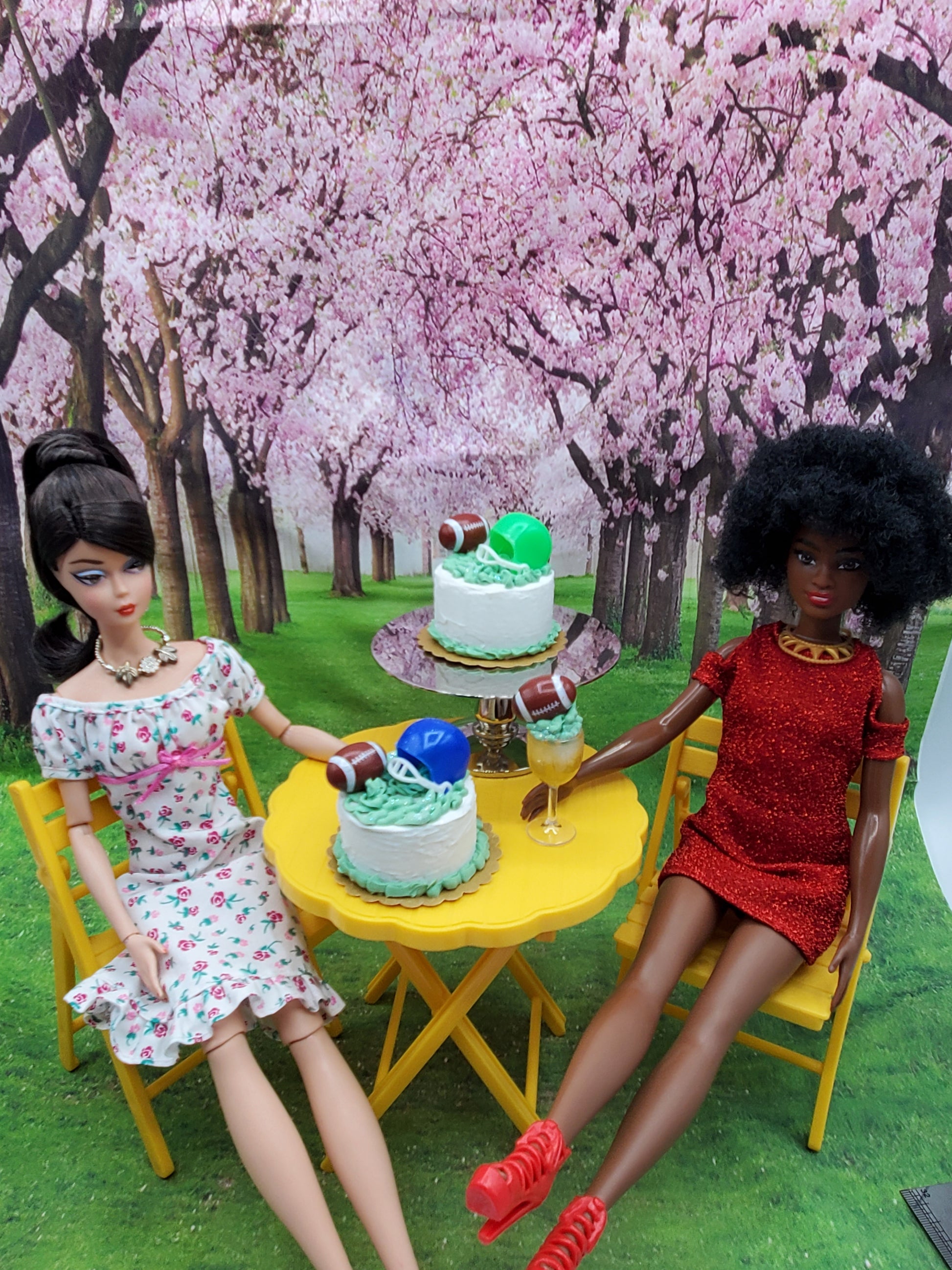 Miniature football cake desserts on yellow table with two dolls seated under cherry blossom trees