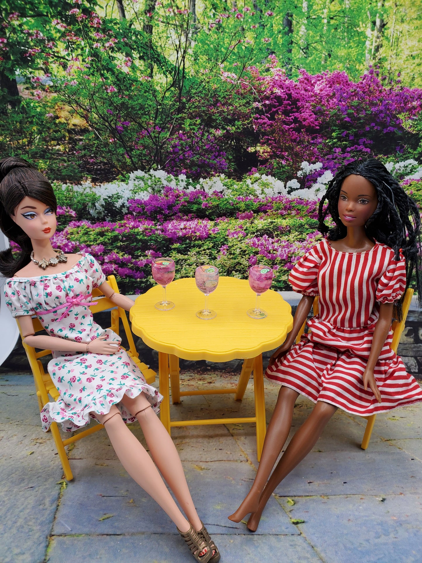 Two dolls sitting at yellow table with three miniature pink lime doll drink glasses in garden setting