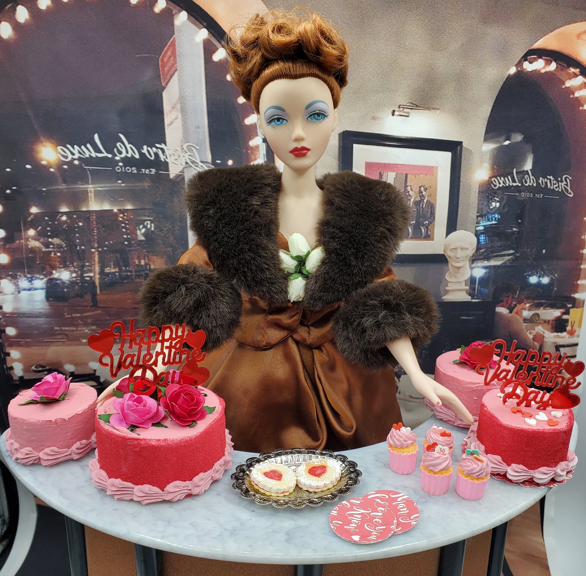 Chef Gina's miniature Valentine's Day cake set with pink cakes and cupcakes displayed with a vintage doll wearing a brown fur-trimmed coat