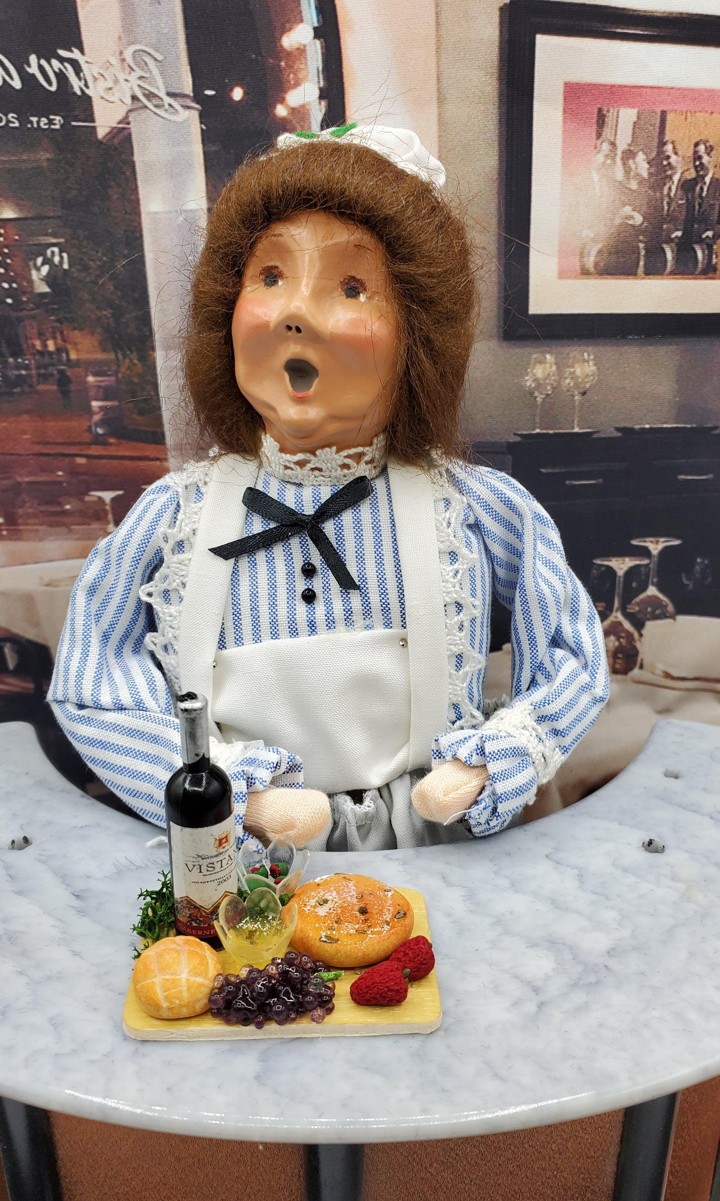 Miniature wine cheese tray with grapes, cheese, and crackers on a table next to a doll dressed as a chef