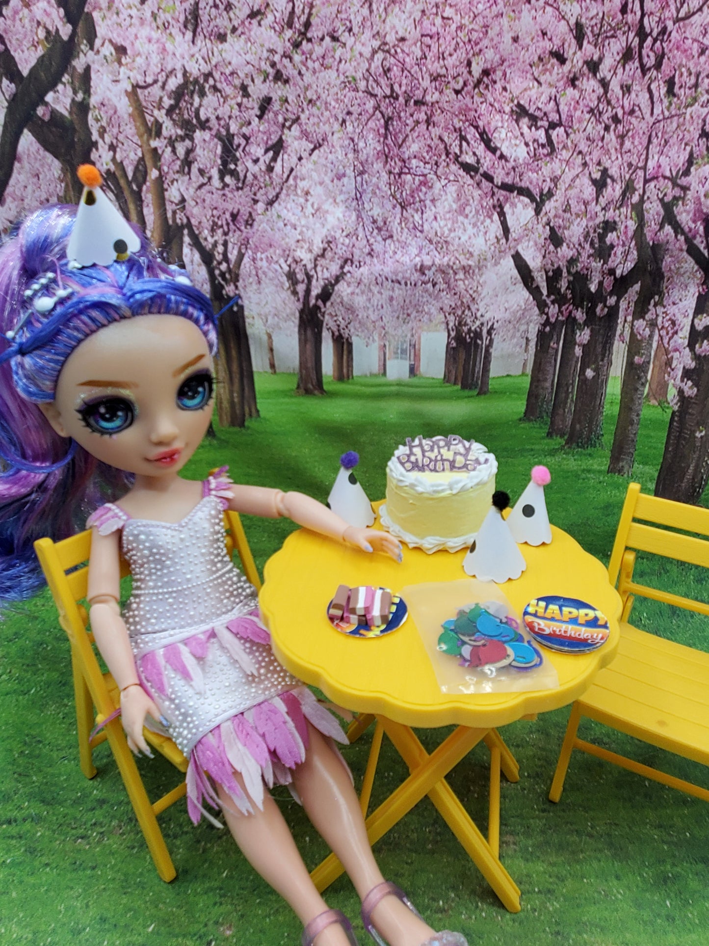 Pink flamingo doll party set featuring a doll with purple hair sitting at a yellow table with birthday cake and party hats