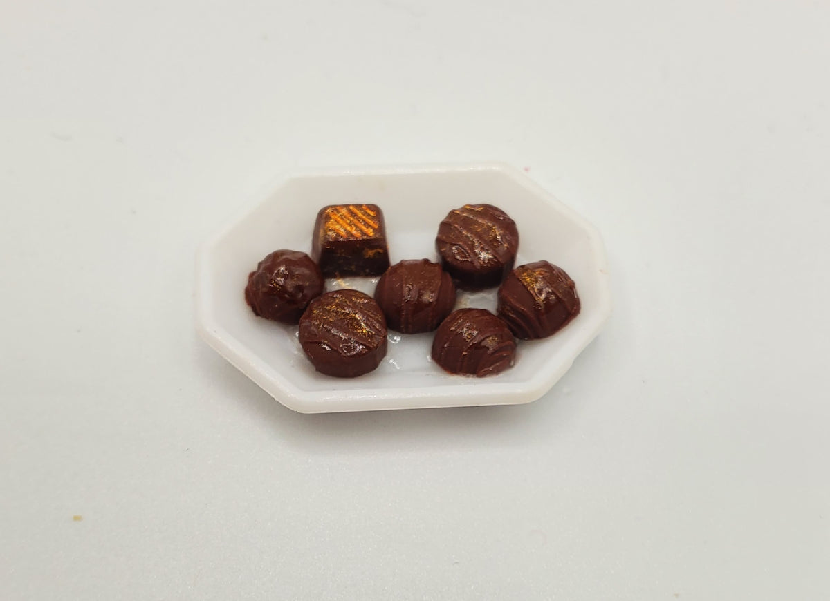 Six miniature chocolate candies, including several milk chocolate round pieces and one dark chocolate square with gold leaf detailing, arranged on a small white octagonal dish against a plain white background.