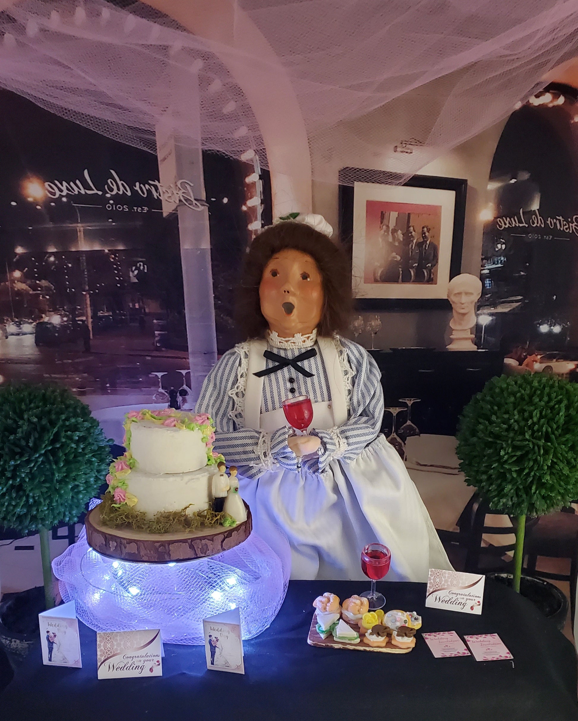 Doll in a striped dress with a cake and wine glasses on a table, with a blurred indoor background.