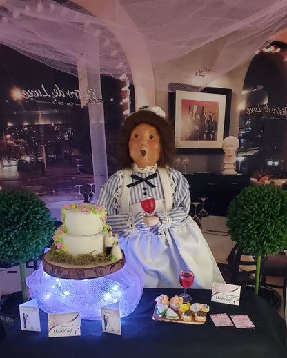Doll in a striped dress with a cake and wine glasses on a table, with a blurred indoor background.