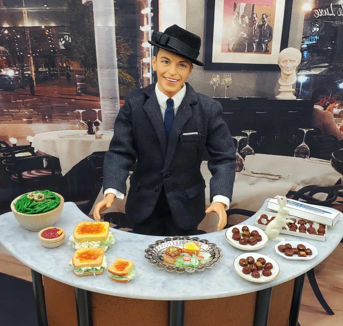 A doll dressed as a waiter in a dark pinstripe suit, white shirt, and black fedora hat stands behind a dining table laden with miniature food items in a restaurant setting.