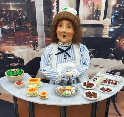 A vintage ceramic doll dressed as a nurse stands behind a curved table laden with miniature food items, set against a blurred restaurant interior background.