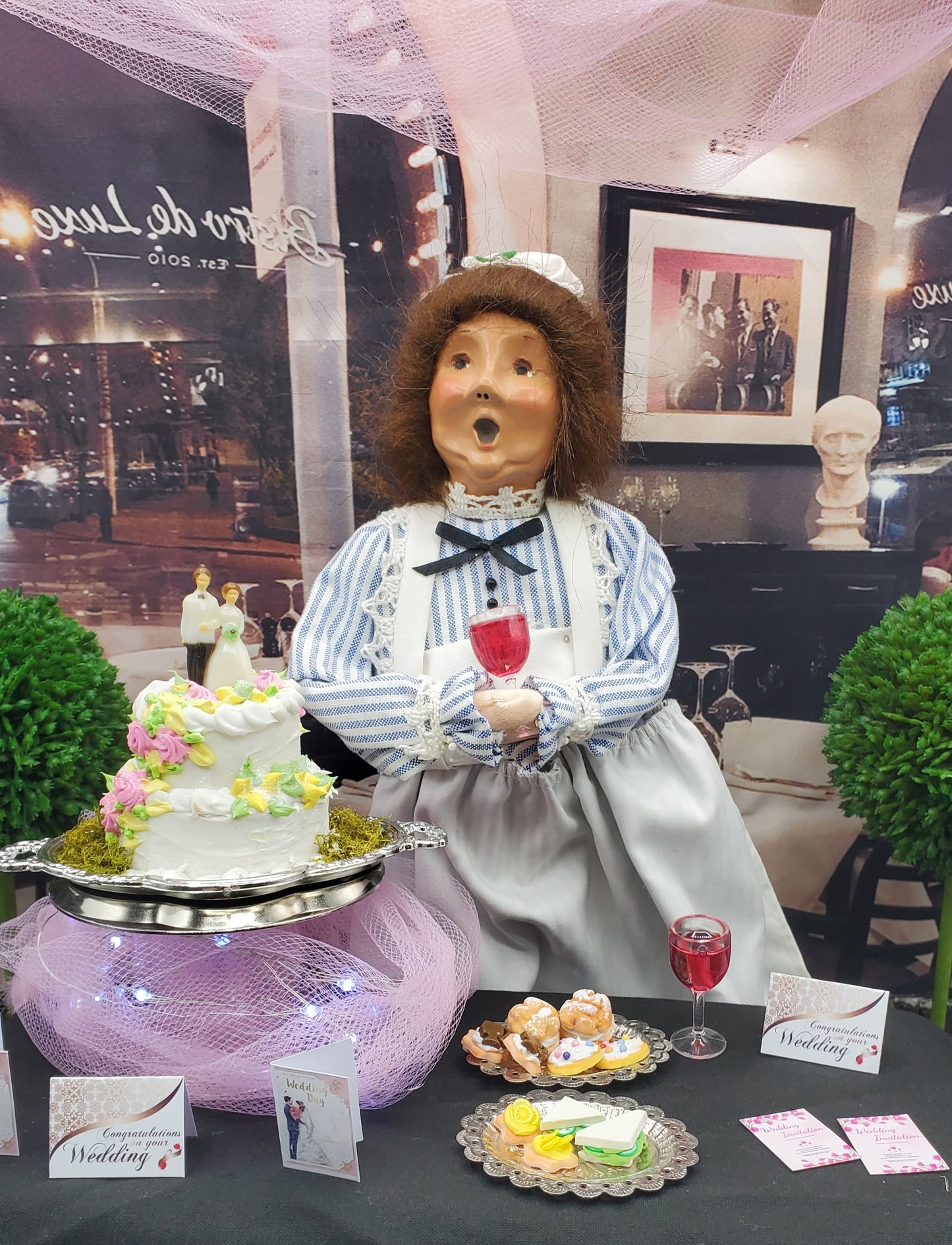 Display of a figurine with a cake and other items on a table, with a cityscape background.