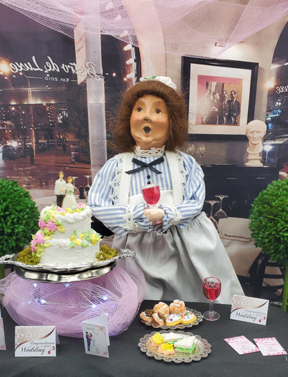 Display of a figurine with a cake and other items on a table, with a cityscape background.