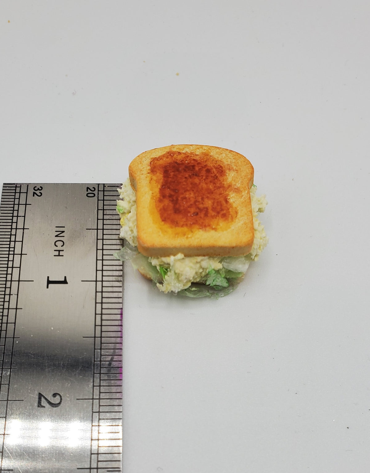A miniature sandwich with a toasted golden-brown top slice, white filling, and green lettuce, shown next to a metal ruler for scale on a plain white background.