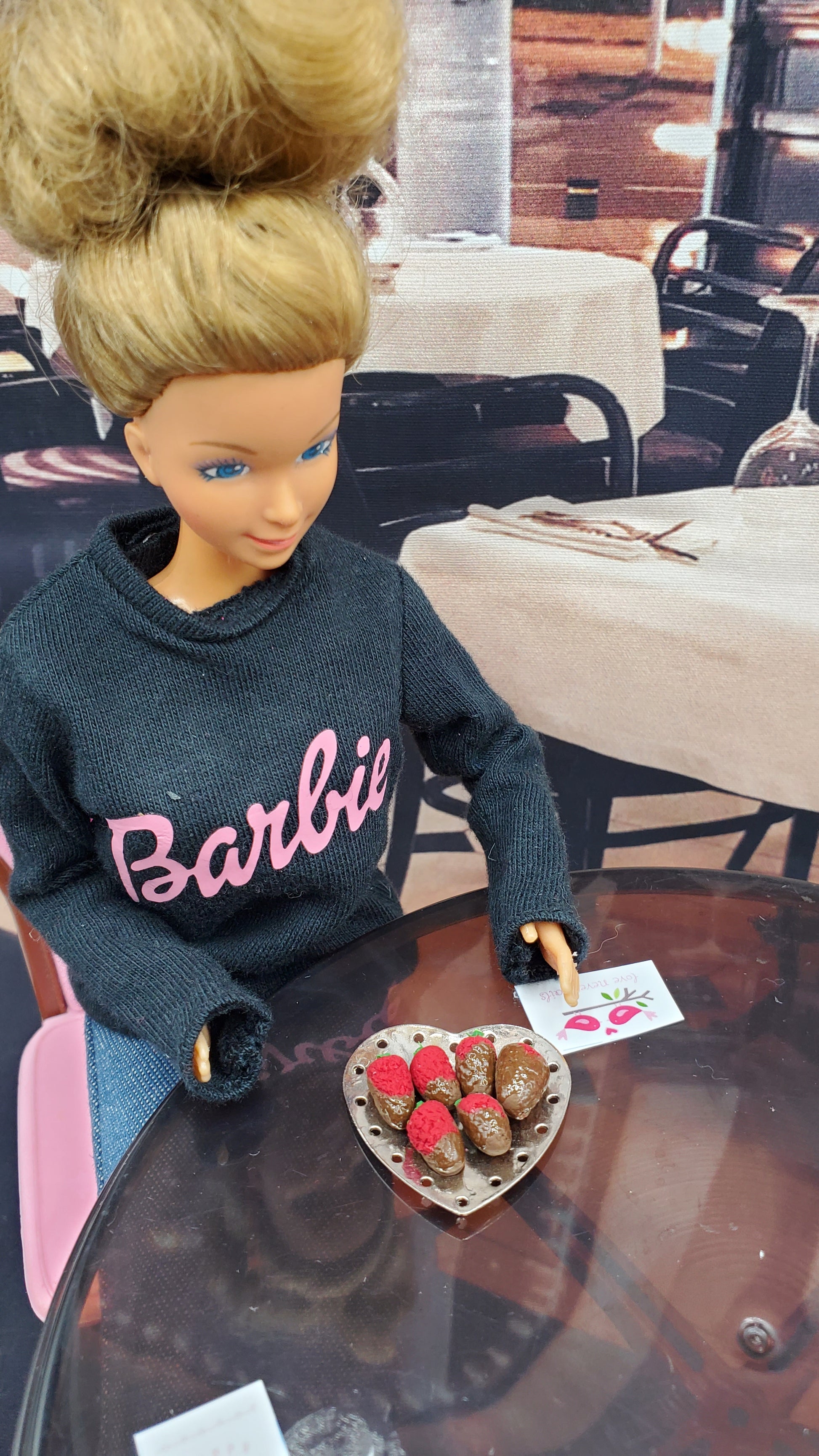 Barbie doll wearing a black outfit with 'Barbie' written on it, sitting on a chair.