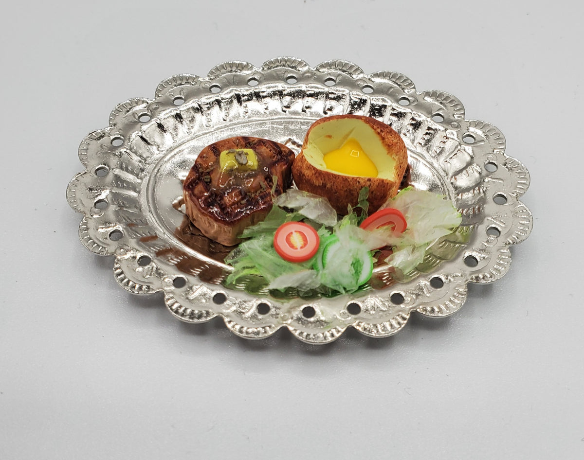 A decorative silver metal serving platter with a scalloped edge, holding a cooked food dish with a halved egg, two small browned pieces, and a small salad garnish on a light gray background.