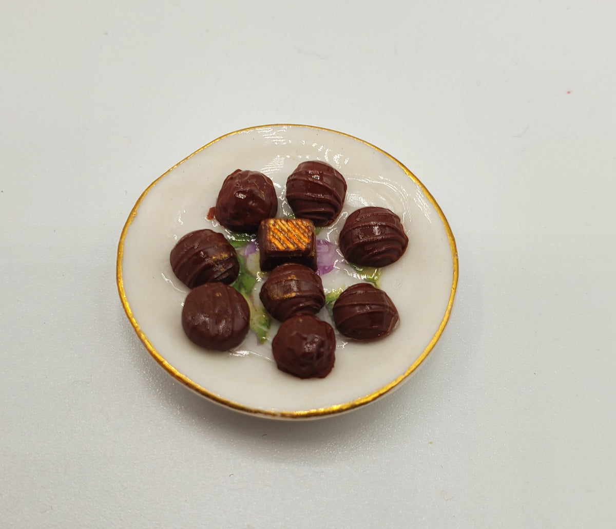 A miniature ceramic plate with a gold rim, holding several small dark brown chocolate-like candies and a small gold-patterned square piece, set against a plain light background.