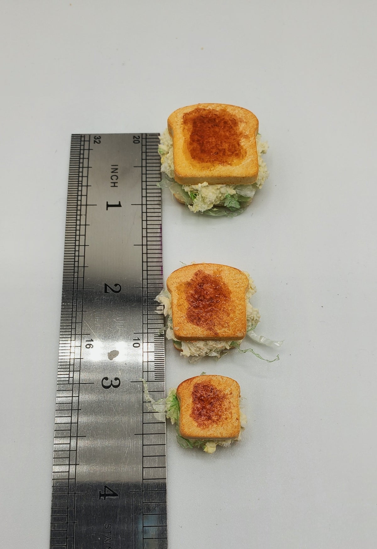 Three miniature sandwich slices with toasted bread and green filling arranged vertically next to a metal ruler on a white background.