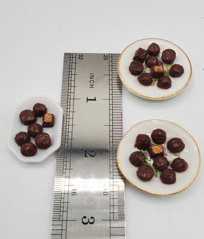 Three miniature white plates with gold trim, each holding several tiny chocolate-dusted candies, arranged around a metal ruler on a white background to show scale.