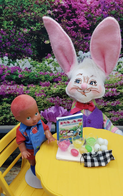 Fashion doll egg decorating kit featuring a small doll and a bunny with decorating supplies on a yellow table outdoors