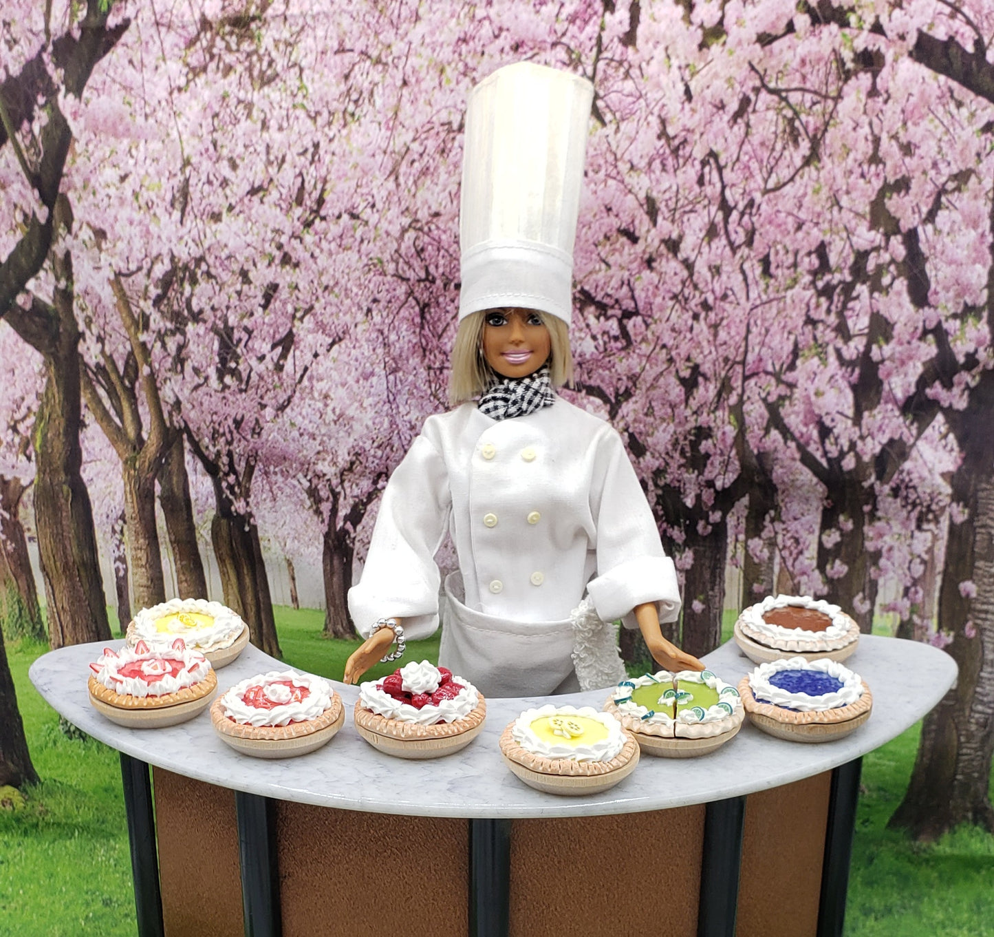 Chef doll presenting an array of miniature banana cream pie and other decorated pies in a floral setting