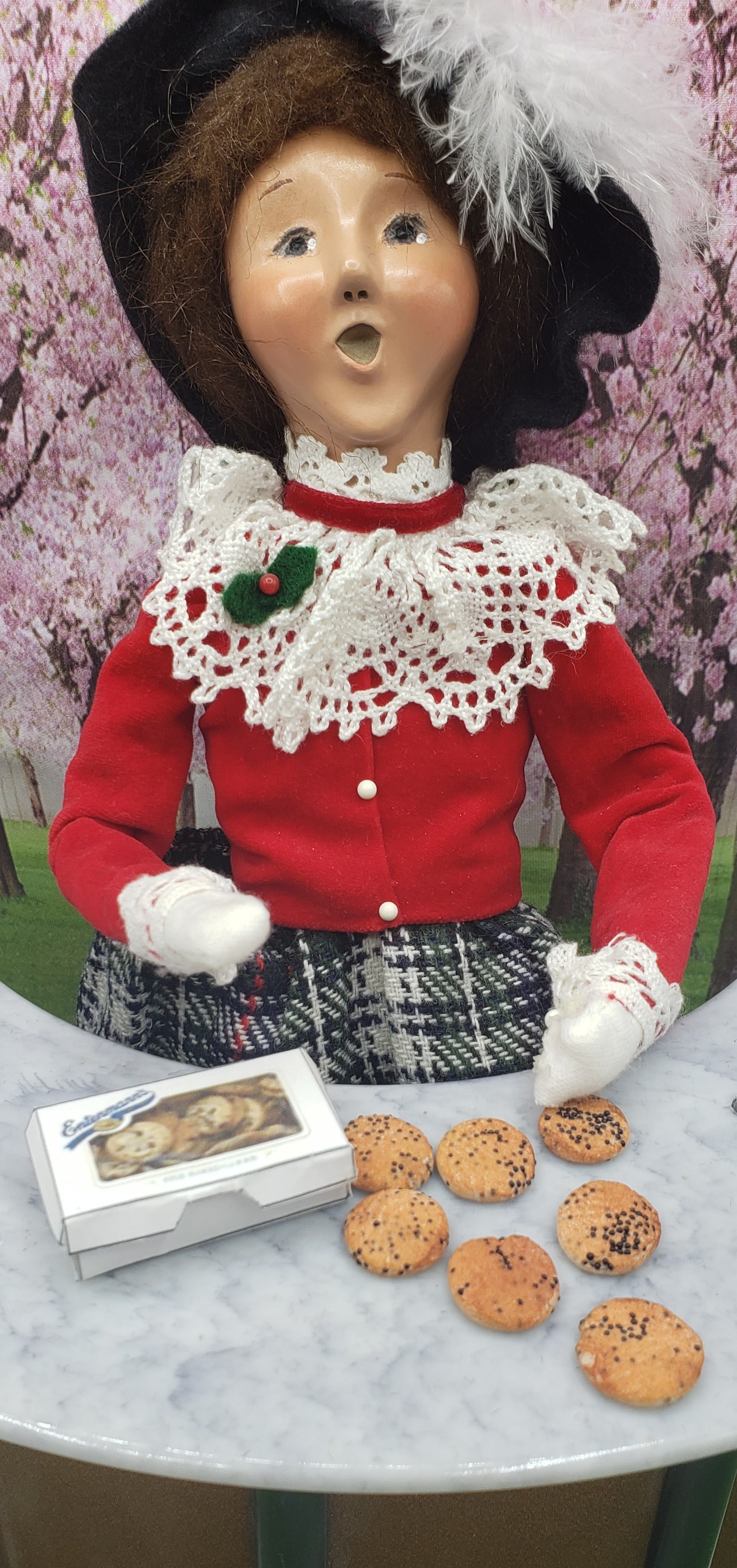 Vintage doll dressed in red and lace holding miniature chocolate chip cookies with cookie box on table