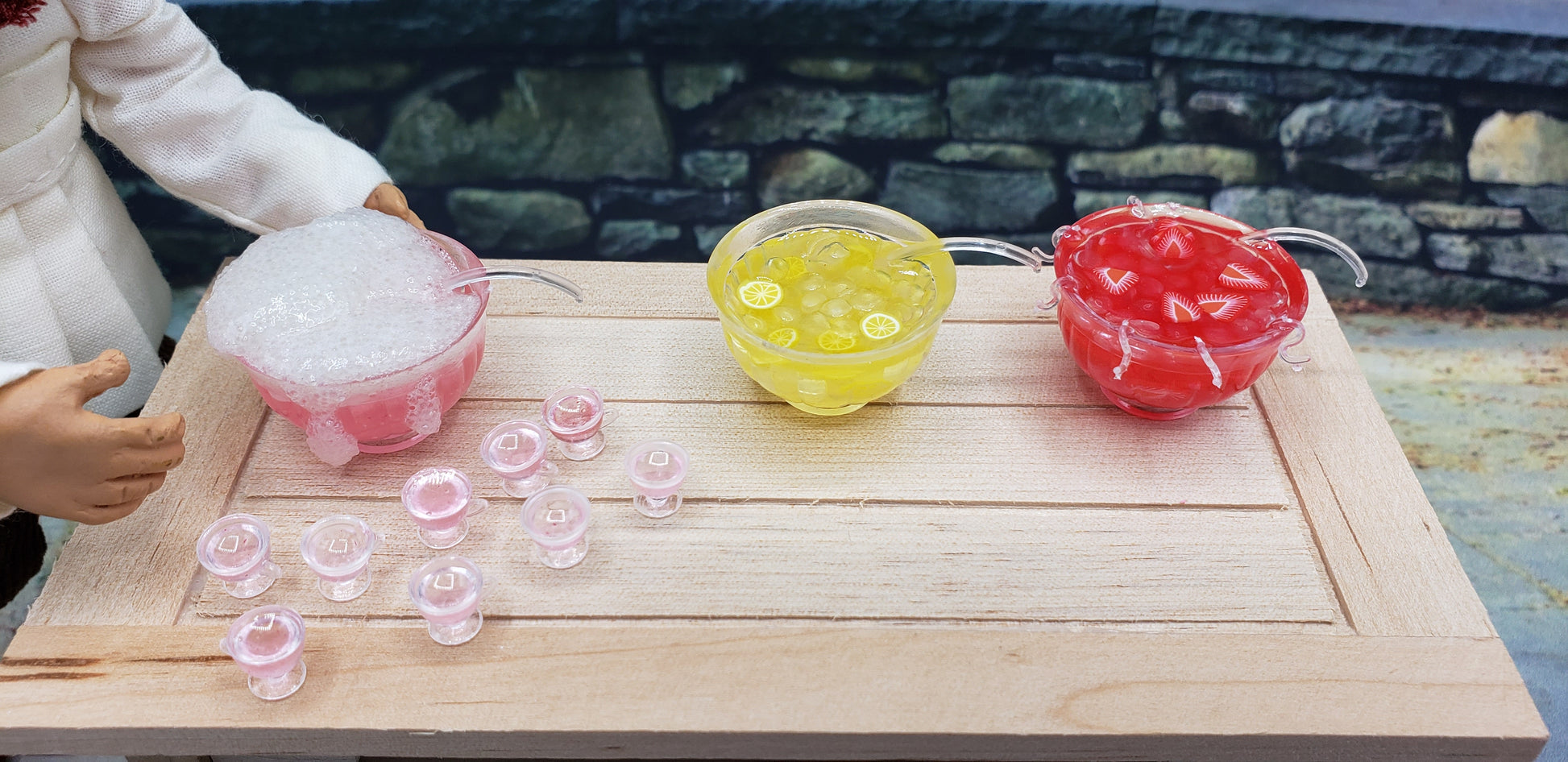 Miniature dollhouse party punch set with red, yellow, and pink bowls and tiny pink punch cups on a wooden table