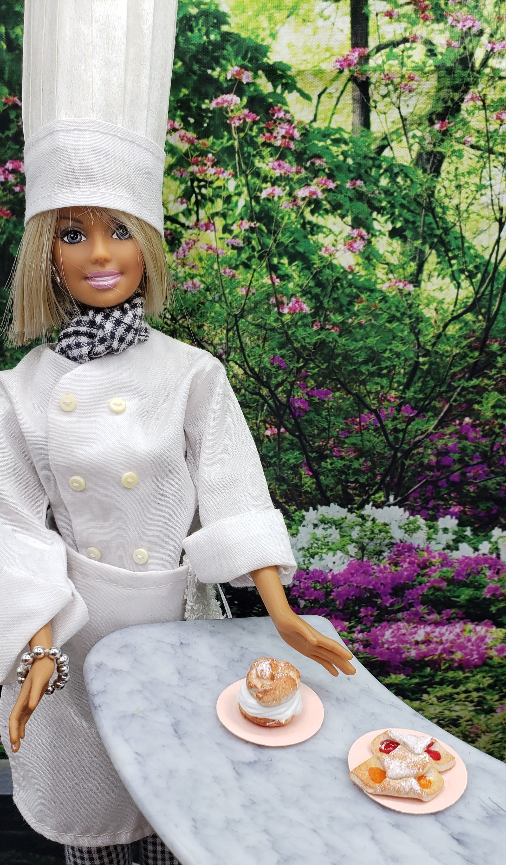 Chef Gina doll presenting miniature koloche doll food on a marble table in a floral garden setting