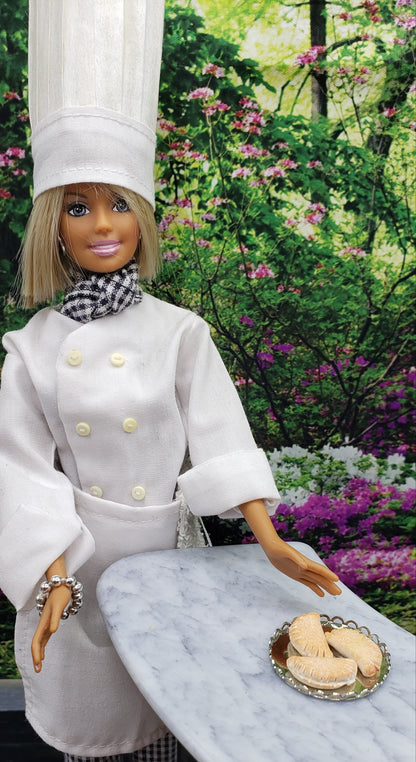 Chef doll presenting a plate of miniature pierogi for dolls on a marble table in a floral garden